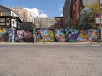 a wall with different colorful artwork on it in a city with buildings around and the street has a bike path going under it