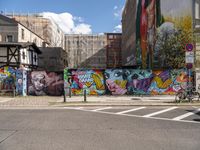 a wall with different colorful artwork on it in a city with buildings around and the street has a bike path going under it