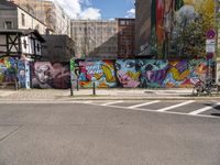 a wall with different colorful artwork on it in a city with buildings around and the street has a bike path going under it