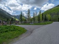 road in the mountains with grass and trees and a fence surrounding it on a bright day