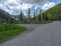 road in the mountains with grass and trees and a fence surrounding it on a bright day