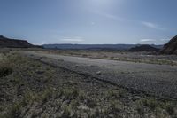 an empty road surrounded by dirt and grass with the sun reflecting over the mountains in the distance