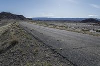 an empty road surrounded by dirt and grass with the sun reflecting over the mountains in the distance