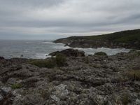 a rocky shore is pictured next to the ocean and an airplane flies overhead in a cloudy sky