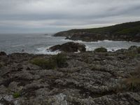 a rocky shore is pictured next to the ocean and an airplane flies overhead in a cloudy sky