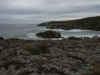 a rocky shore is pictured next to the ocean and an airplane flies overhead in a cloudy sky