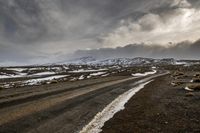 a dirt road under a gray sky with snow and rocks beside it in the foreground