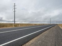 an empty long road is shown on this cloudy day with power lines across it and telephone poles to the side