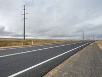 an empty long road is shown on this cloudy day with power lines across it and telephone poles to the side