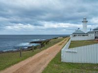 a dirt road that is next to a white lighthouse by the water on a cloudy day