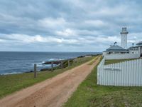 a dirt road that is next to a white lighthouse by the water on a cloudy day