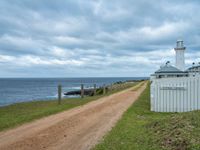 a dirt road that is next to a white lighthouse by the water on a cloudy day