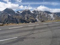 an asphalt road in front of snow capped mountains and a cloud covered blue sky is seen