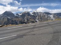 an asphalt road in front of snow capped mountains and a cloud covered blue sky is seen