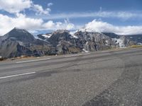 an asphalt road in front of snow capped mountains and a cloud covered blue sky is seen