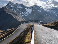 an asphalt road with snow capped mountains in the background behind it is a white fence that leads the end to the roadway with two people