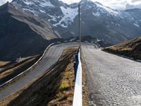 an asphalt road with snow capped mountains in the background behind it is a white fence that leads the end to the roadway with two people