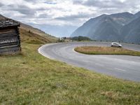 a van driving down an empty country road surrounded by mountains and grassy fields under grey skies