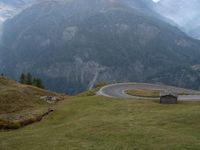 a wooden house stands next to a winding mountain road with a curved road and mountains behind it