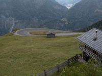 a wooden house stands next to a winding mountain road with a curved road and mountains behind it