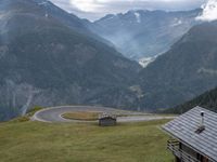 a wooden house stands next to a winding mountain road with a curved road and mountains behind it