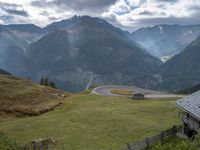 a wooden house stands next to a winding mountain road with a curved road and mountains behind it