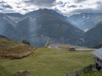 a wooden house stands next to a winding mountain road with a curved road and mountains behind it