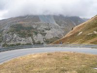 the motorcycle rides on the mountain side of a curved mountain road, with a low cloud covering the top