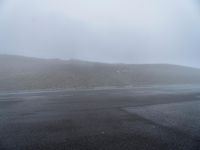 an empty highway with several cars on it in the distance as a hillside is covered by a lot of cloud