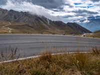 a curved road near many mountains on a cloudy day with clouds in the sky and people walking on the sidewalks