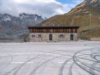 a building with some windows and snow covered mountains behind it, along with tire marks in the ground