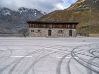 a building with some windows and snow covered mountains behind it, along with tire marks in the ground