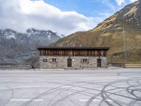 a building with some windows and snow covered mountains behind it, along with tire marks in the ground