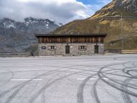 a building with some windows and snow covered mountains behind it, along with tire marks in the ground