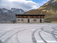 a building with some windows and snow covered mountains behind it, along with tire marks in the ground