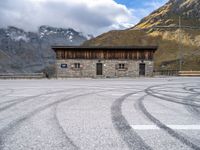 a building with some windows and snow covered mountains behind it, along with tire marks in the ground