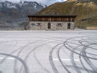 a building with some windows and snow covered mountains behind it, along with tire marks in the ground