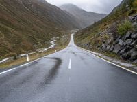 a wet road winds through the mountains to the right of the camera with a stop sign beside it