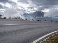 a paved road with mountains behind it under a cloudy sky and clouds above it at the top of a mountain range