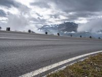 a paved road with mountains behind it under a cloudy sky and clouds above it at the top of a mountain range