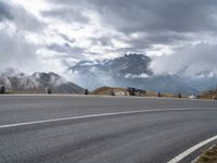 a paved road with mountains behind it under a cloudy sky and clouds above it at the top of a mountain range