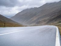 a lone road stretches through a wide valley with hills in the background below and some clouds above it