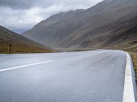 a lone road stretches through a wide valley with hills in the background below and some clouds above it