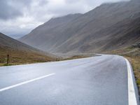 a lone road stretches through a wide valley with hills in the background below and some clouds above it