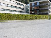 a person skateboards by a block of building with a hill behind them and an evergreen - lined sidewalk and bushes around