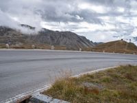 the street with signs is empty by the mountains and grass and rocks as clouds are rising behind it