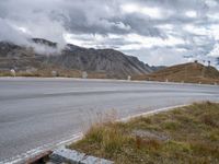 the street with signs is empty by the mountains and grass and rocks as clouds are rising behind it