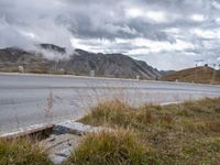 the street with signs is empty by the mountains and grass and rocks as clouds are rising behind it