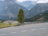 a street with a curve in the middle of mountains, next to a road that looks down from a cliff, and a lone tree on top