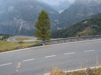 a street with a curve in the middle of mountains, next to a road that looks down from a cliff, and a lone tree on top